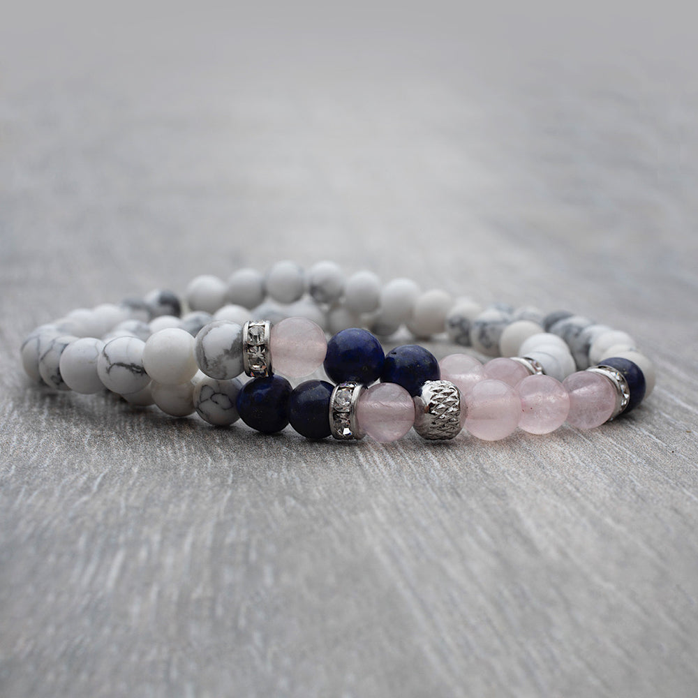 
                  
                    Duo de bracelets élastiques faits avec des pierres fines howlite, quartz rose et lapis lazuli déposé sur une table de bois.
                  
                
