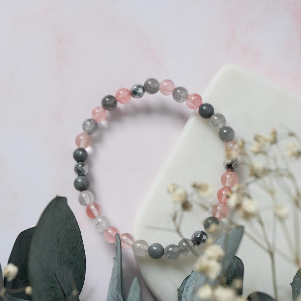 
                  
                    Bracelet de pierres de quartz rose, cerise et fumé ainsi que d’hématite déposé sur un livre et fleurs séchées
                  
                